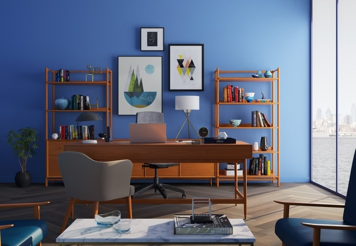 Landscape styled picture of an office space. There is a big panoramic window on the right wall of the picture. A wooden desk sits in the center of the room with matching wooden bookshelves behind it. Each bookshelf is lined with mid-century modern decor. There is a waist high plant in the left corner of the room. The entire room is painted a deep ocean blue. 
