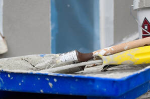 A blue paint tray lays on the floor, it is filled with two paint brushes and one paint roller. Both the roller and the brushes are soaked in white paint. The roller has a yellow handle. 