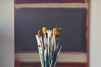 at least 10 paint brushes are standing vertically in a clear cup. Behind the brushes is a wall that is painted purple with red and white stripes. The brushes look wet like they have been recently cleaned. 
