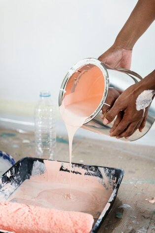 A personas hands are in a close up view pouring a silver gallon sized bucket of paint into a pan to be rolled onto a wall. The paint being poured is pink, and the floor underneath is layered with cardboard to protect it. 