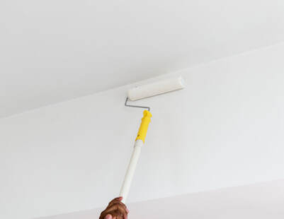 A man reaches to the ceiling with a pole connected to a paint roller. The paint roller is covered in white paint.